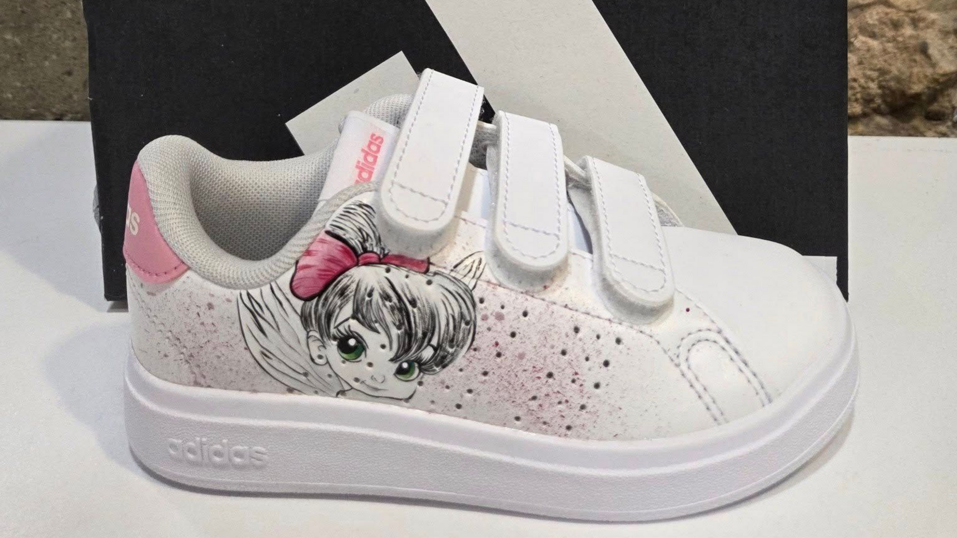Chaussure blanche Adidas pour tout-petit avec des accents roses, un design de personnage et des sangles velcro.