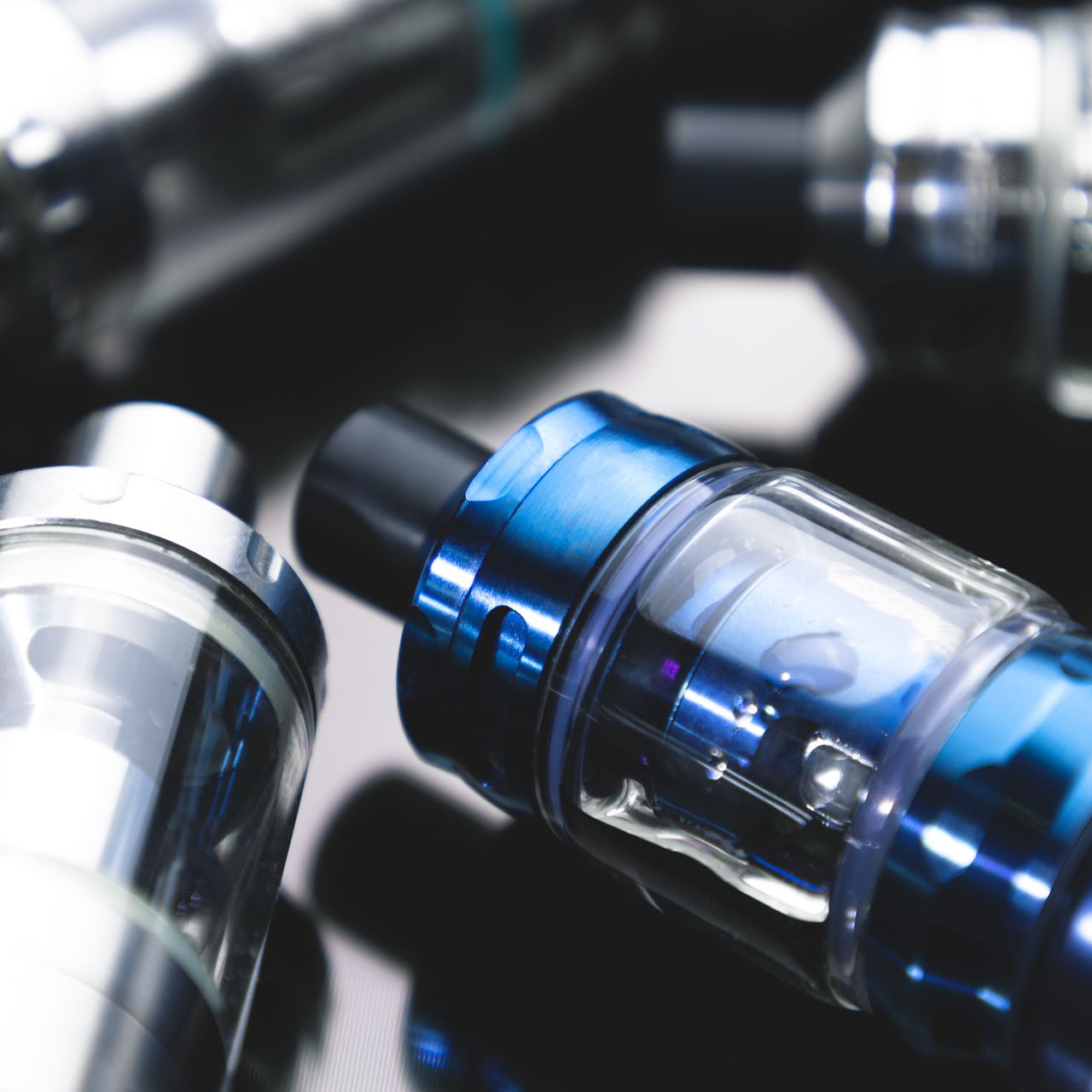 Gros plan de plusieurs stylos vape brillants sur une surface sombre réfléchissante, certains avec des accents bleus.