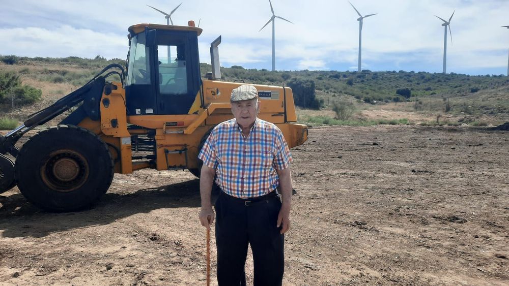 Un hombre está parado frente a un tractor amarillo en un campo con molinos de viento en el fondo.