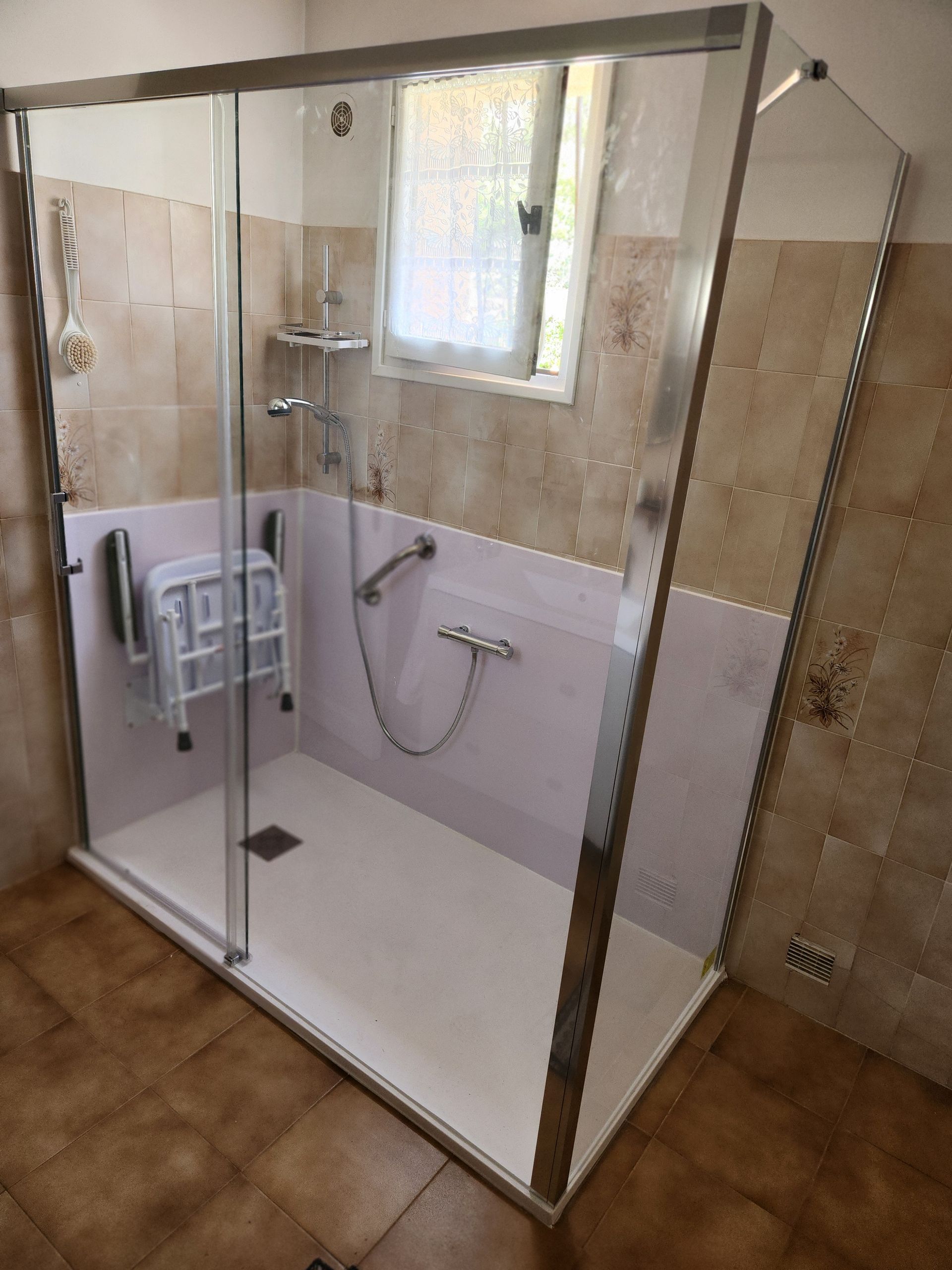 Douche à l'italienne accessible avec paroi en verre, murs blancs, carrelage beige et barre d'appui près de la fenêtre.