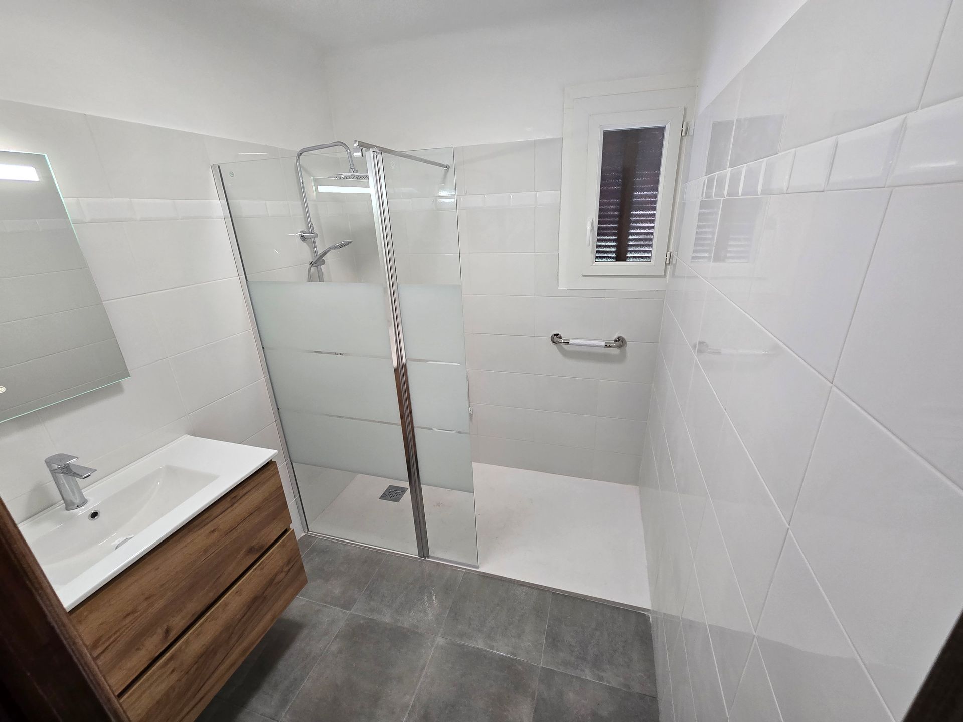 Salle de bains moderne avec douche en verre, murs carrelés blancs, meuble-lavabo en bois et carrelage gris au sol.