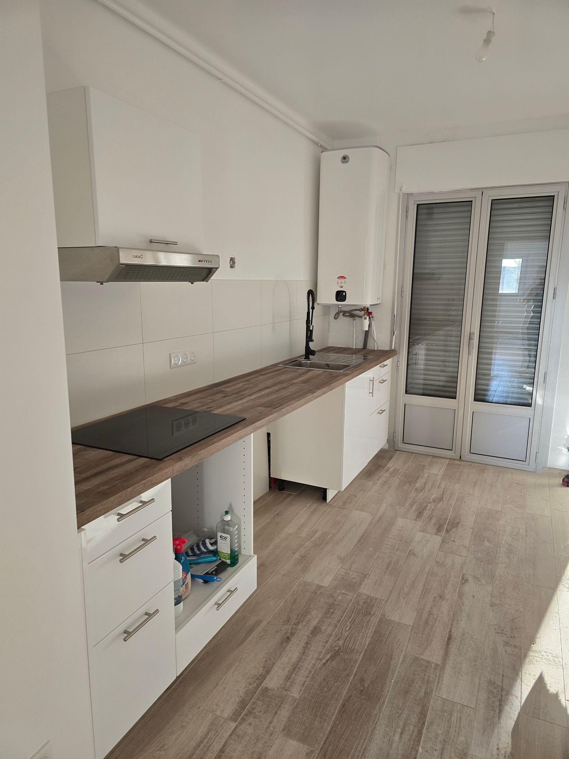 Cuisine vide avec placards blancs, plan de travail en bois et portes vitrées à volets au fond.
