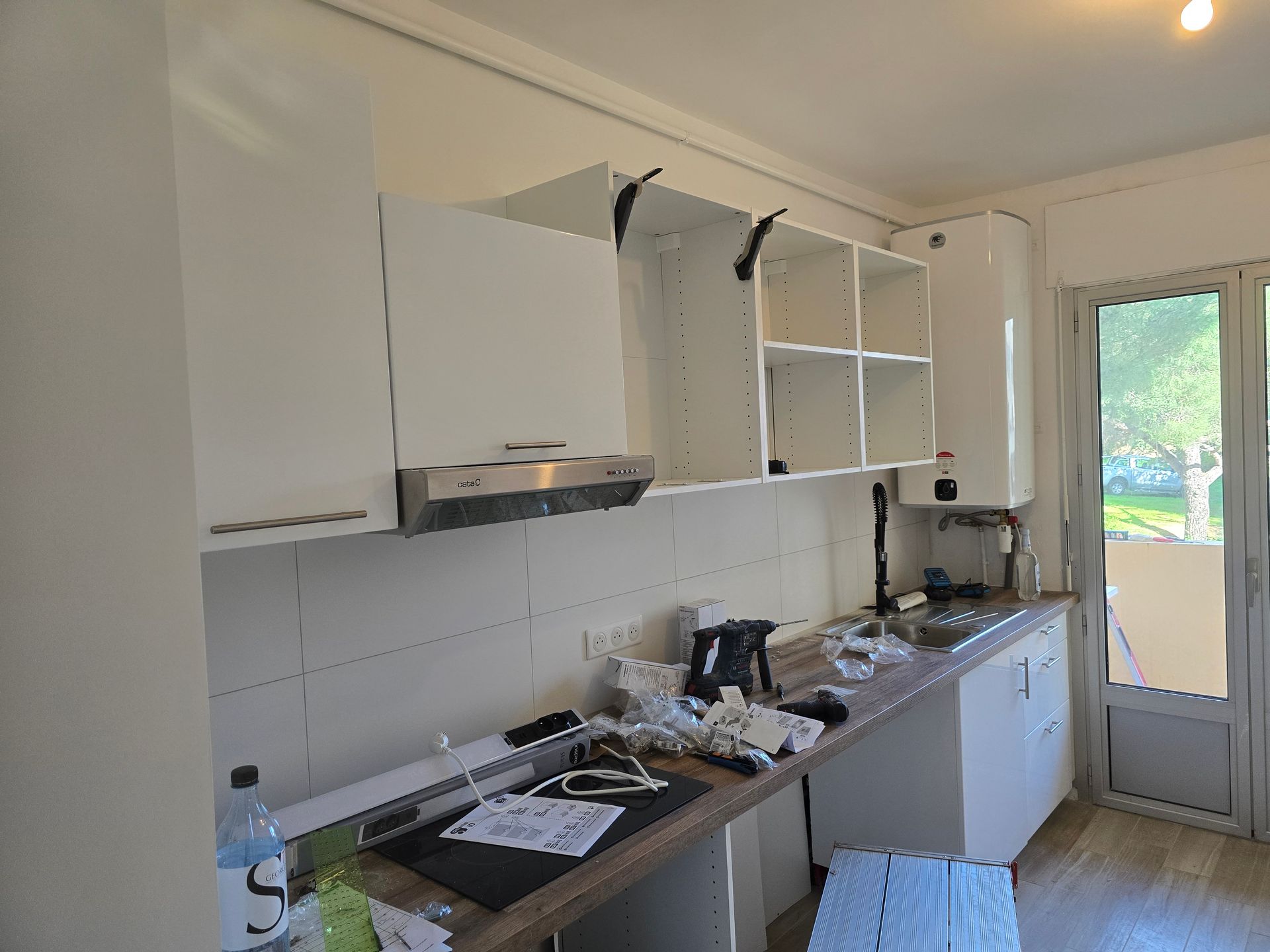 Cuisine en désordre avec des placards blancs, un plan de travail encombré et une porte donnant sur un jardin extérieur.