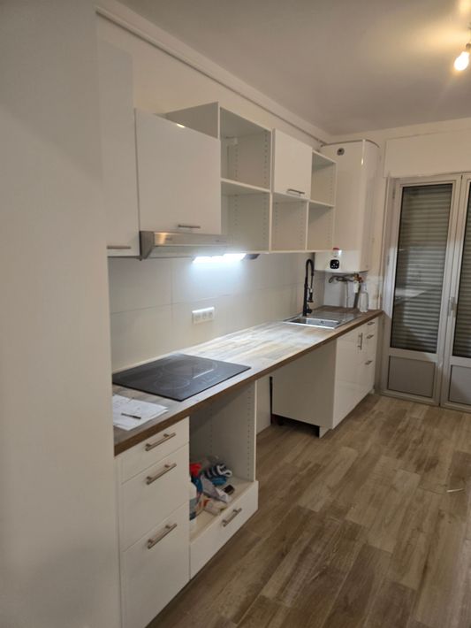 Cuisine moderne blanche en forme de couloir avec parquet, plan de travail, placards, évier, plaque de cuisson et porte-fenêtre donnant sur un balcon.