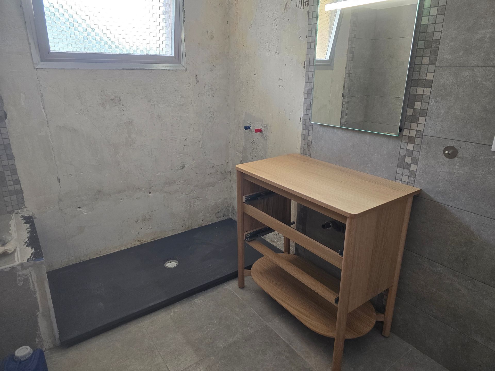 Petite salle de bains avec murs en béton brut, receveur de douche noir et meuble-lavabo en bois sous un miroir.