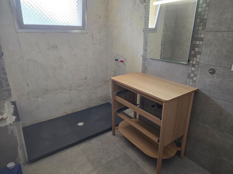 Petite salle de bains avec murs en béton brut, receveur de douche noir et meuble-lavabo en bois sous un miroir.