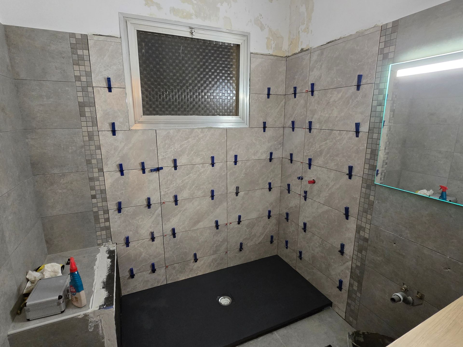 Salle de bains partiellement carrelée avec receveur de douche noir, fenêtre, miroir et accessoires muraux apparents.