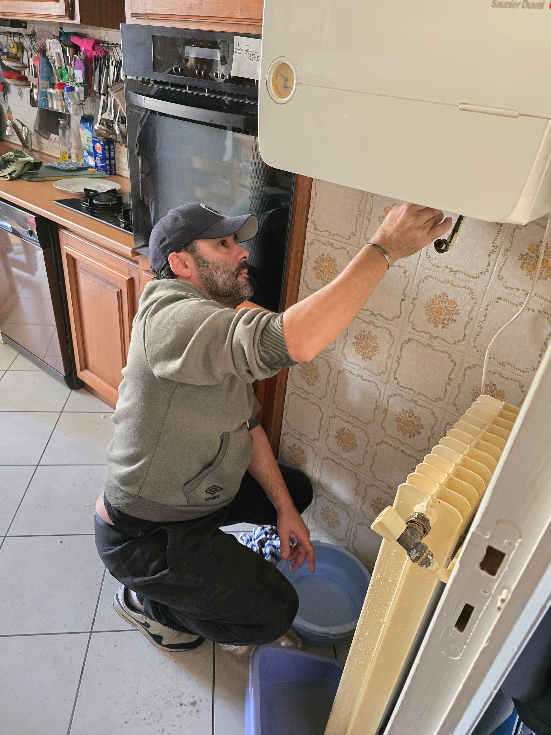 Un technicien est agenouillé dans une cuisine, en train de réparer un appareil électroménager mural au-dessus d'un radiateur.