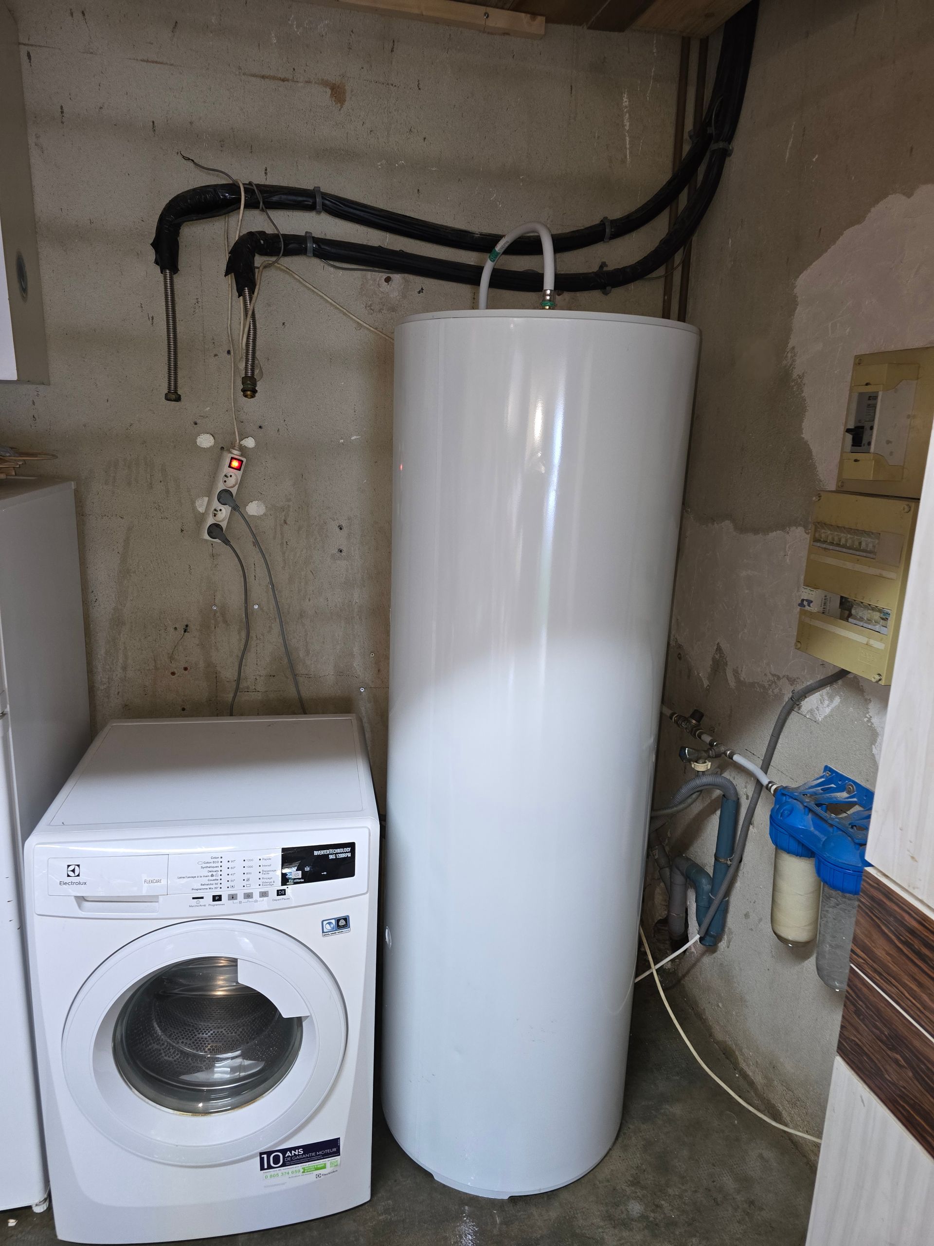 Buanderie avec une machine à laver blanche à côté d'un grand chauffe-eau blanc et des tuyaux apparents au mur