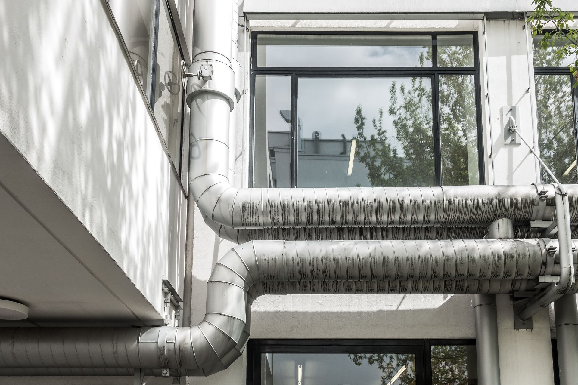 De larges conduits argentés traversent la façade blanche d'un bâtiment, à côté d'une fenêtre et d'arbres.