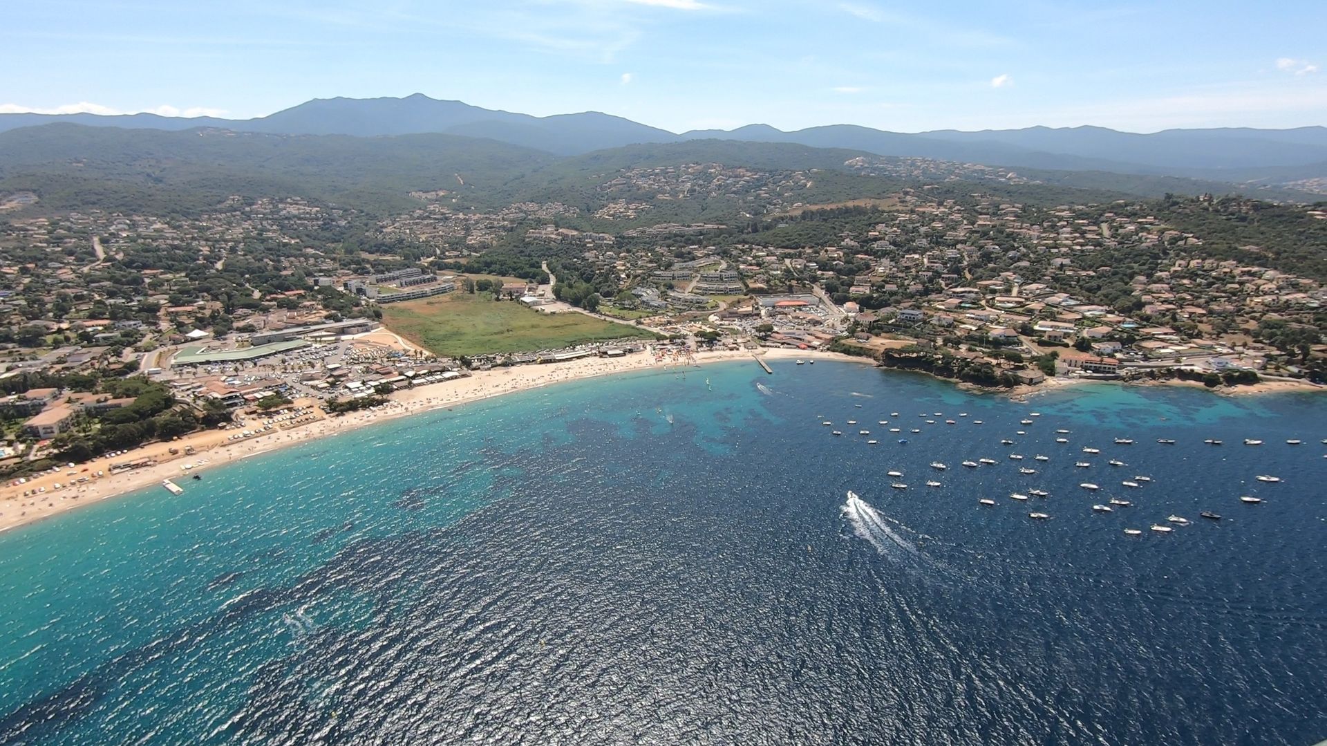 Vue aérienne d'une baie turquoise avec une plage de sable fin, des bateaux et une ville côtière adossée à des collines.