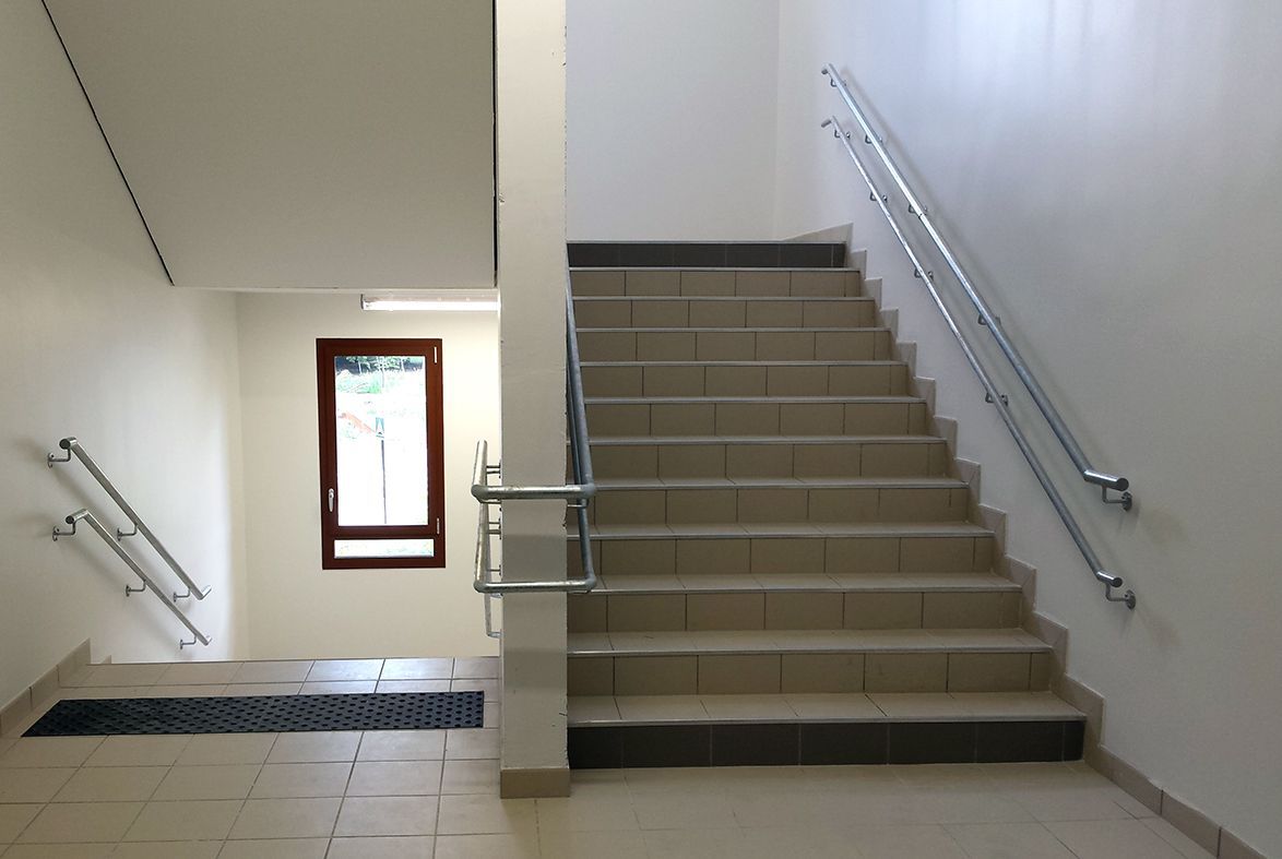 Escalier intérieur avec marches carrelées, rampes métalliques et petite fenêtre donnant sur un couloir lumineux