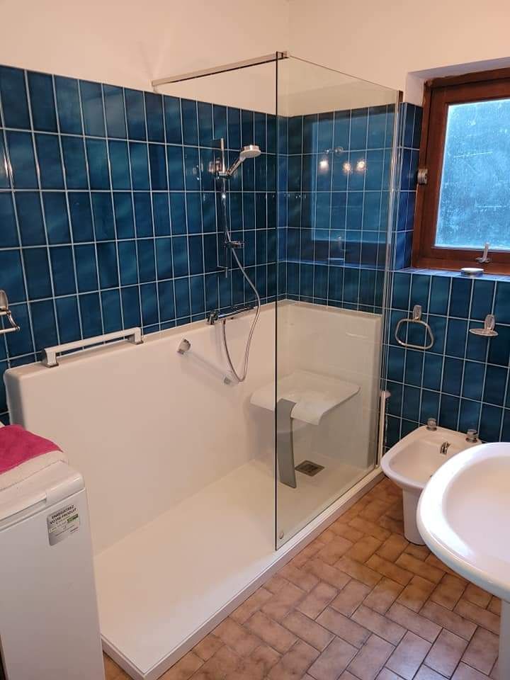 Salle de bains avec murs carrelés bleus, cabine de douche en verre, toilettes, lavabo et fenêtre.