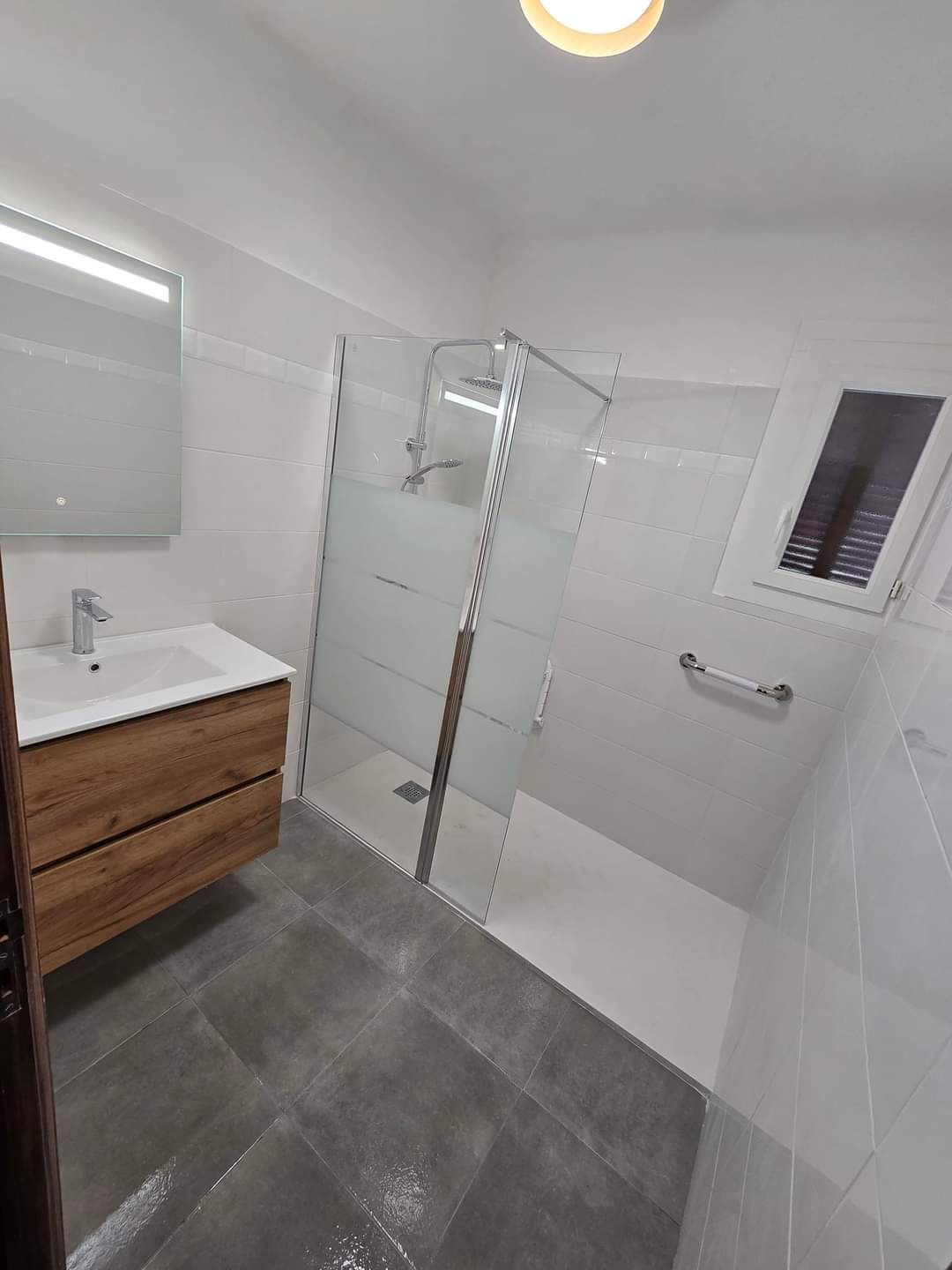 Salle de bains moderne avec douche à l'italienne en verre, meuble-lavabo en bois, carrelage blanc et sol gris.
