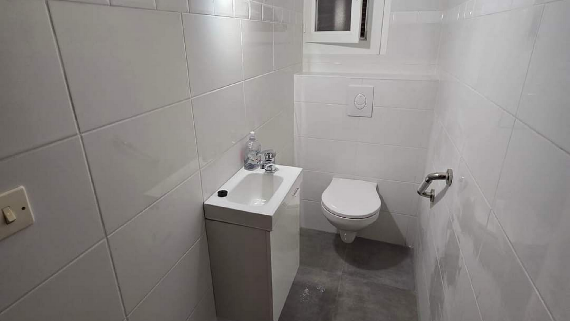 Salle de bains étroite carrelée de blanc avec un petit lavabo, des toilettes et un porte-papier toilette mural.