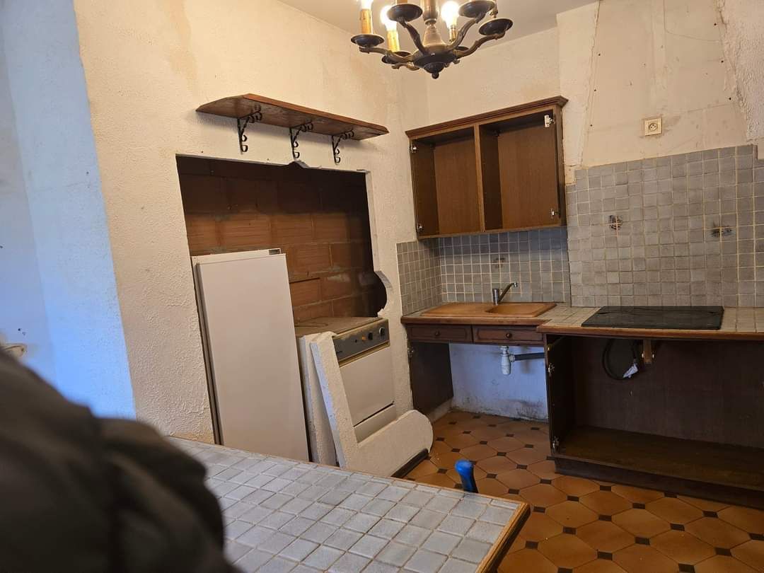 Petite cuisine avec sol carrelé, placards en bois, évier, cuisinière et réfrigérateur sous une fenêtre.