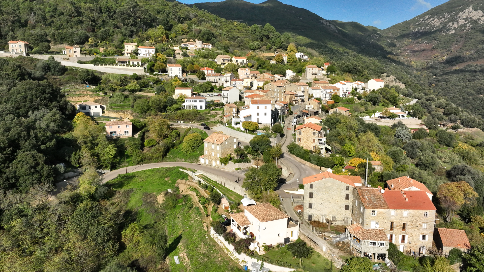 Village à flanc de colline avec des maisons aux toits rouges et des routes sinueuses au milieu de montagnes verdoyantes sous un ciel d'un bleu limpide