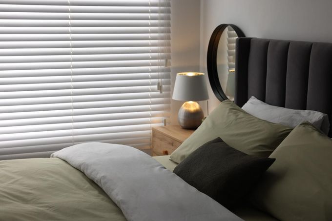 Acogedora habitación con cama, cabecero acolchado gris, lámpara de noche y persianas.