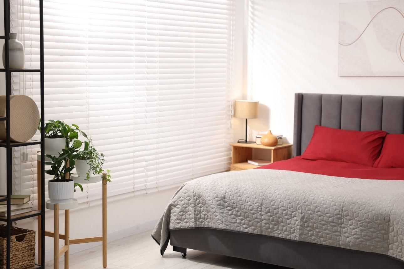 Habitación luminosa con una cama roja, una manta gris, persianas blancas y una pequeña planta sobre un soporte dorado.
