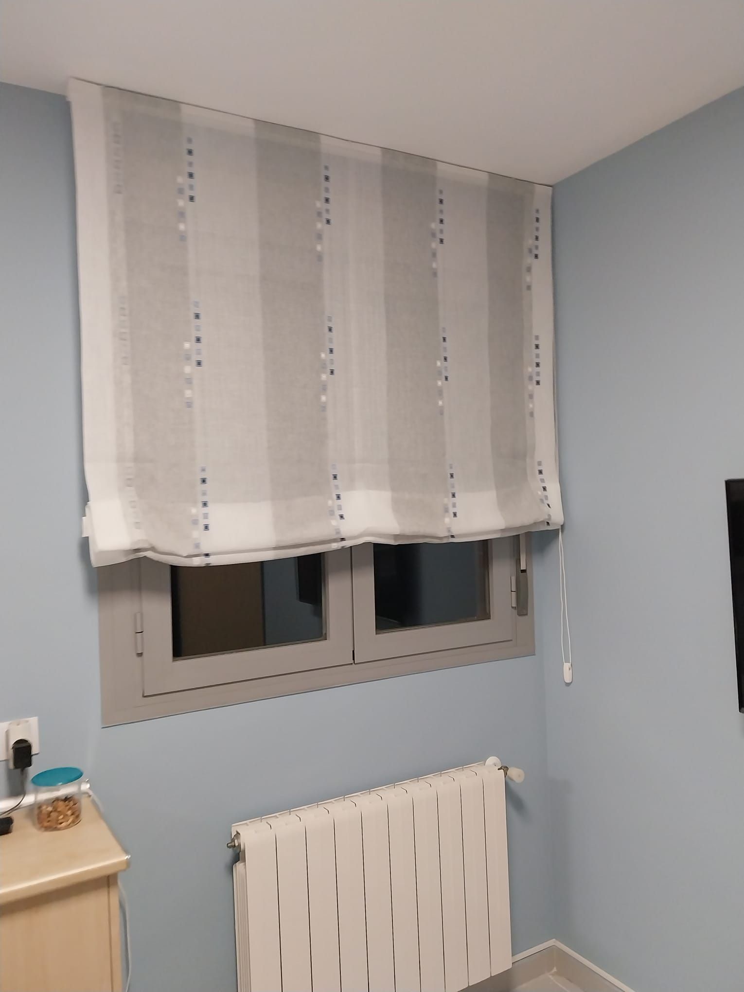 Estor romano blanco estampado sobre una pequeña ventana encima de un radiador blanco en una habitación azul claro.