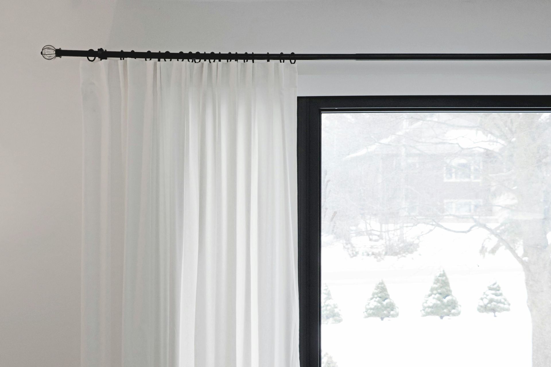 Cortinas blancas junto a una gran ventana con marco negro que muestra una escena exterior nevada.