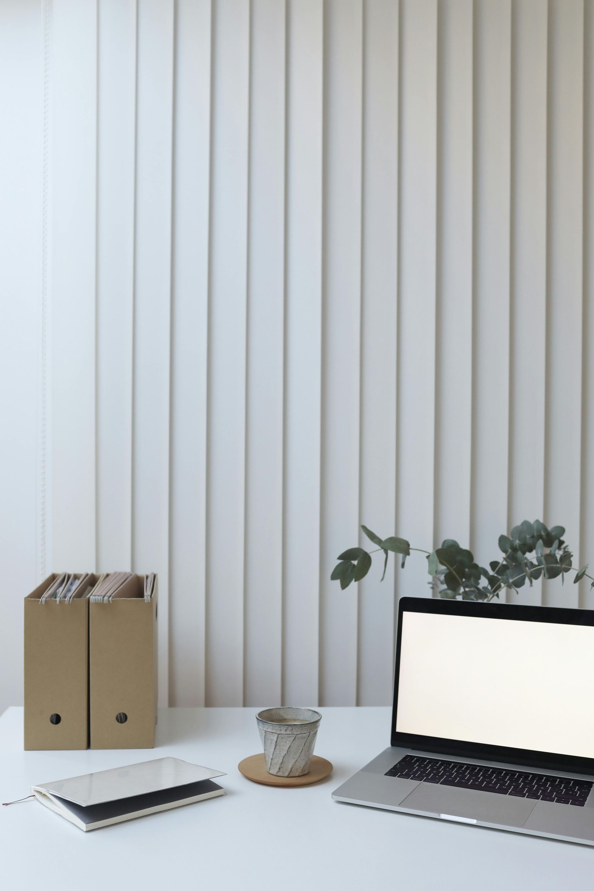 Escritorio blanco minimalista con portátil, cuaderno, portalápices y planta frente a persianas verticales.