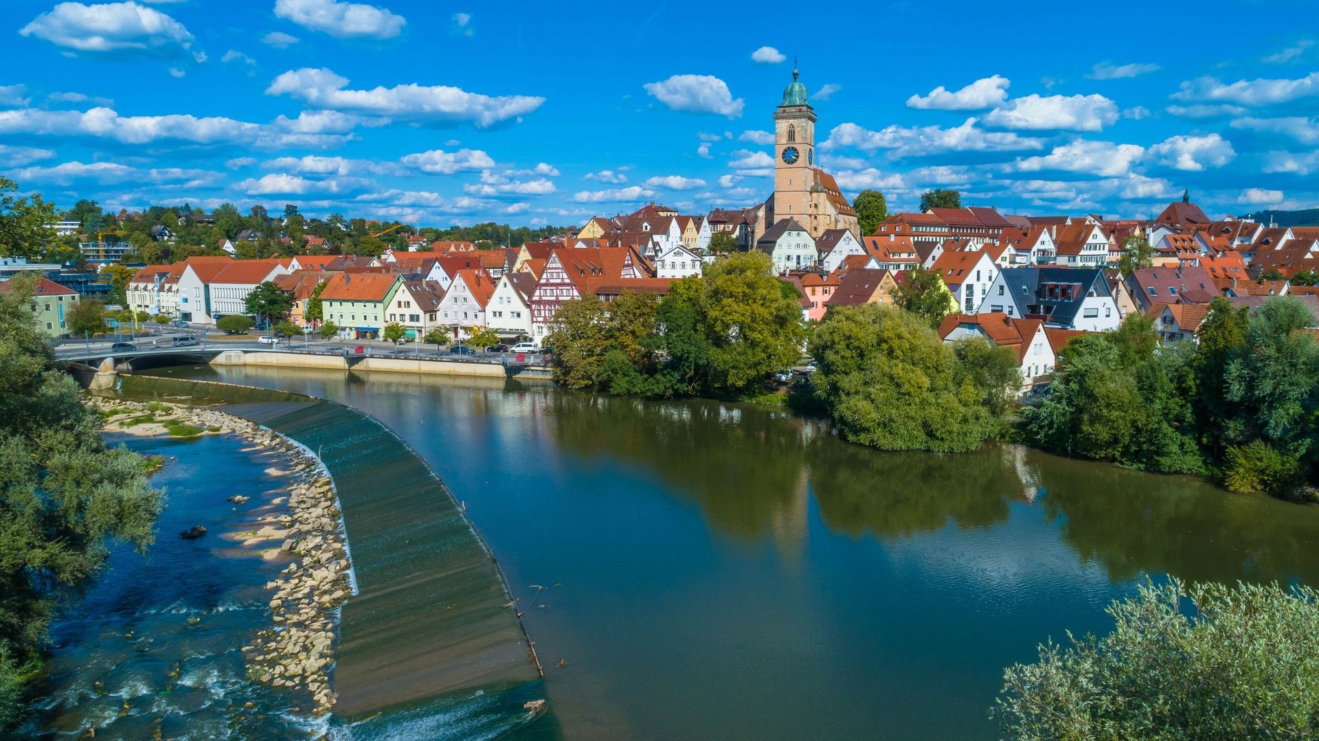 Stadtbild mit Fluss und Kirche unter blauem Himmel. Häuser mit roten Ziegeldächern und grünen Bäumen.