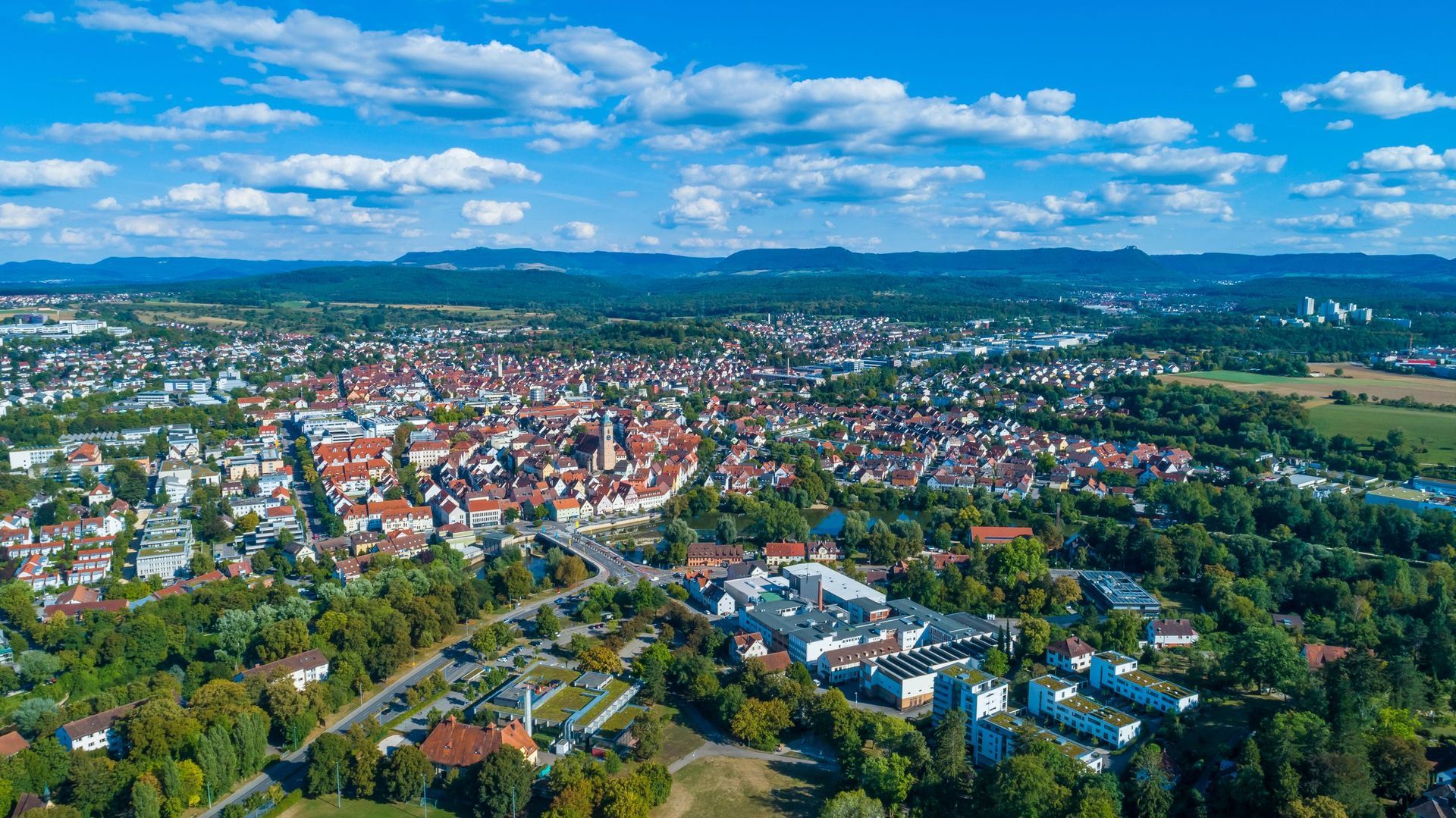 Luftaufnahme einer Stadt mit roten Ziegeldächern, umgeben von grünen Bäumen unter einem blauen Himmel mit Wolken.