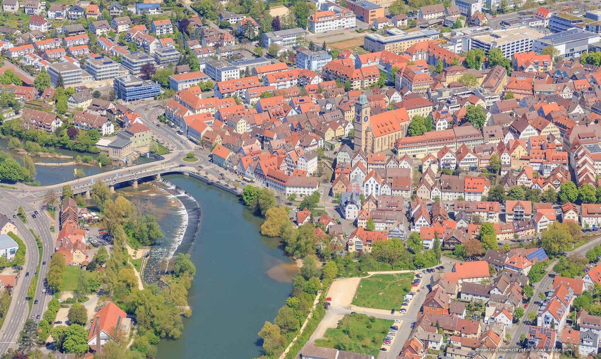 Luftaufnahme einer Stadt mit roten Ziegeldächern, einem Fluss, einer Brücke und einer Kirche. Üppiges Grün umgibt die Gebäude.