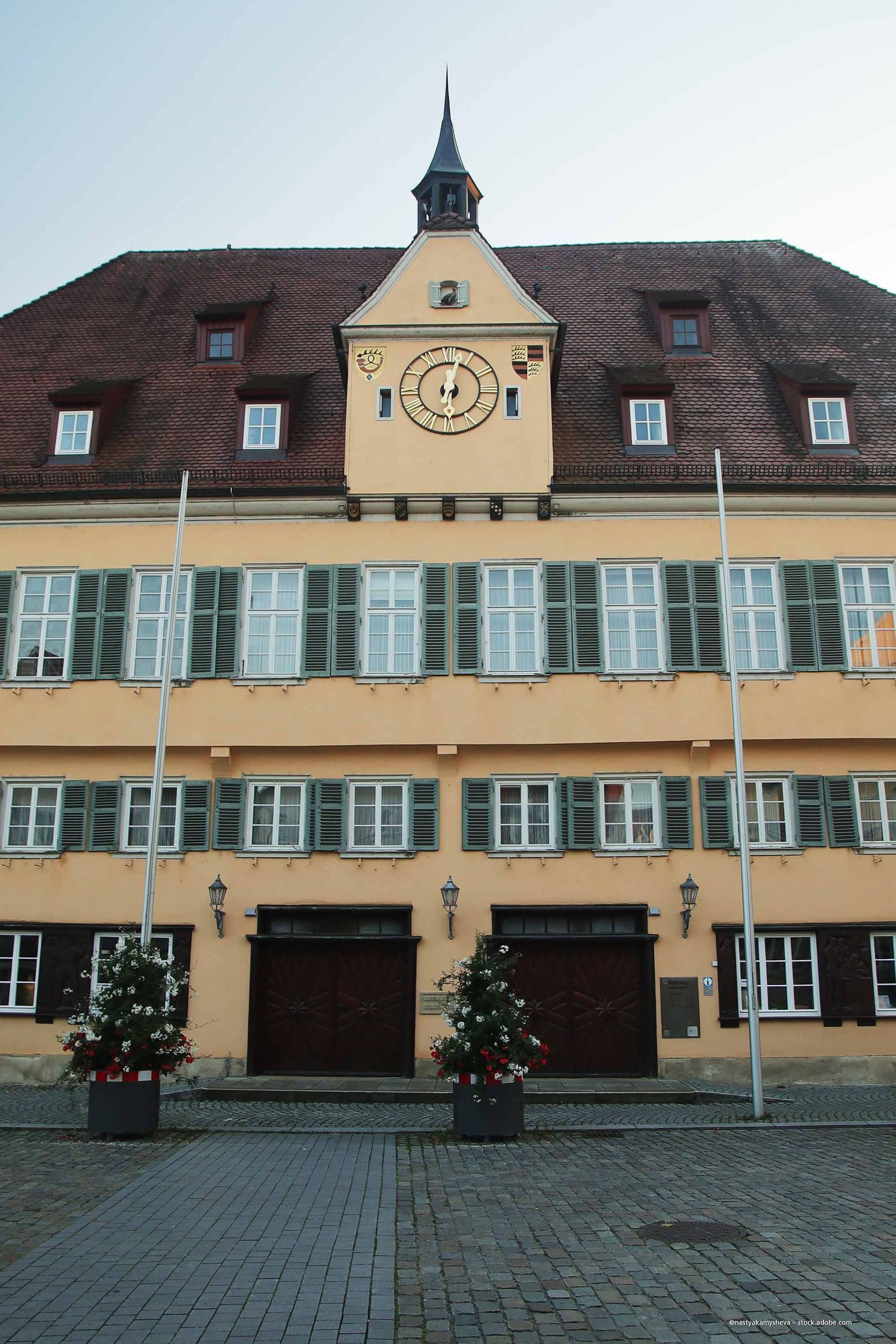 Gelbes Gebäude mit Uhrturm, rotem Dach und grünen Fensterläden. Zwei Bäume flankieren den Eingang.