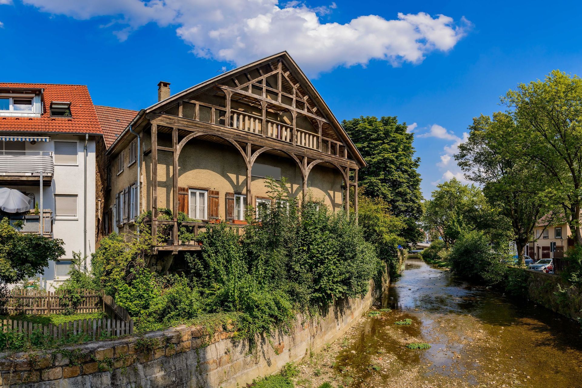 Ein historisches Gebäude mit einem Holzbalkon überblickt einen Bach unter blauem Himmel.