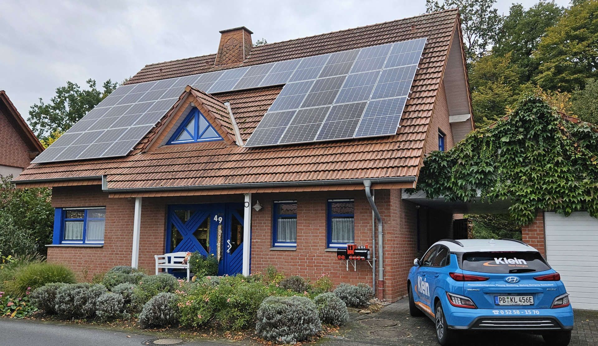 Ein blaues Auto parkt vor einem Backsteinhaus mit Solarzellen auf dem Dach.