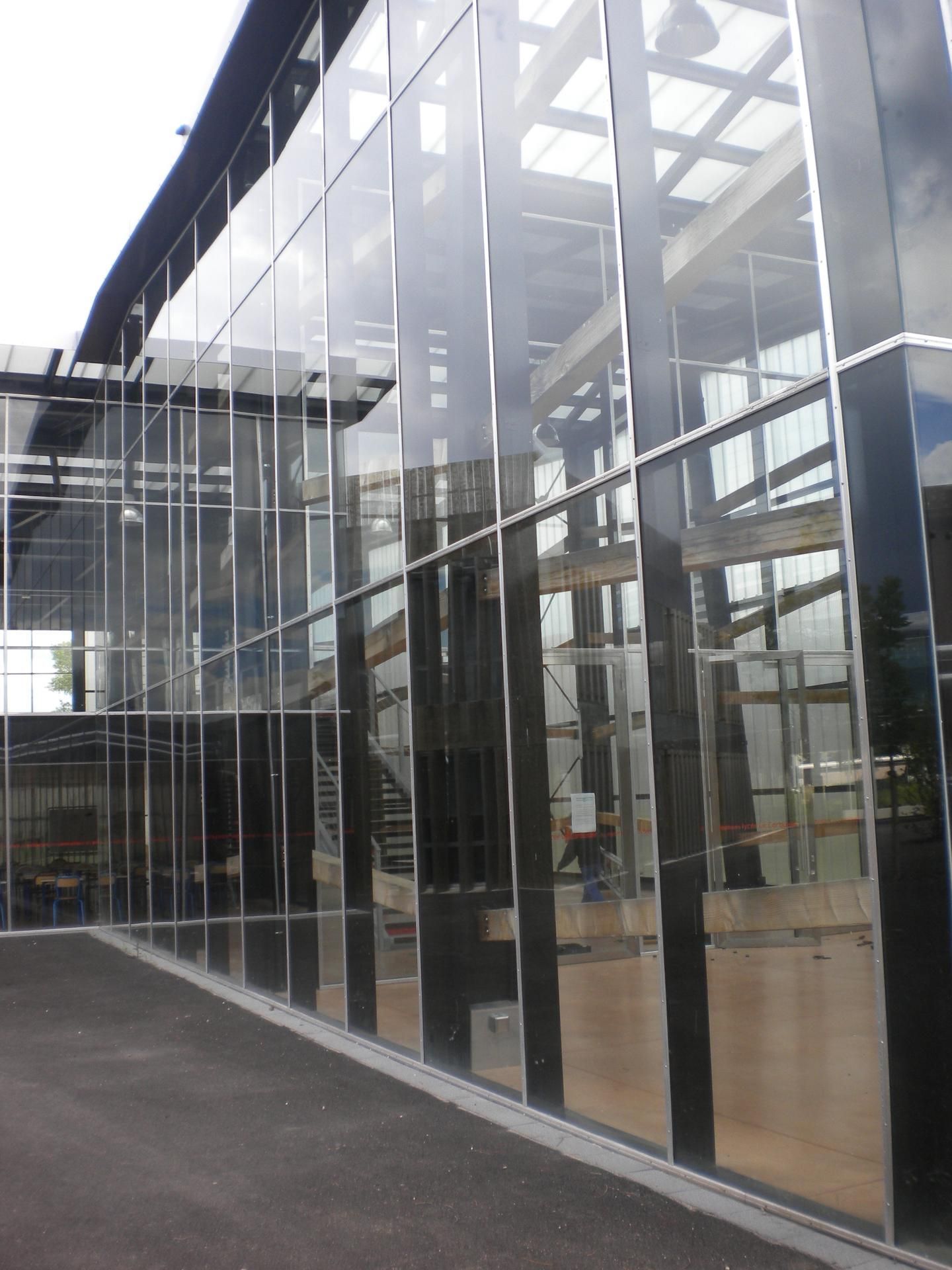 Bâtiment avec fenêtres en verre et entrée couverte, façade grise et blanche, parking au premier plan.
