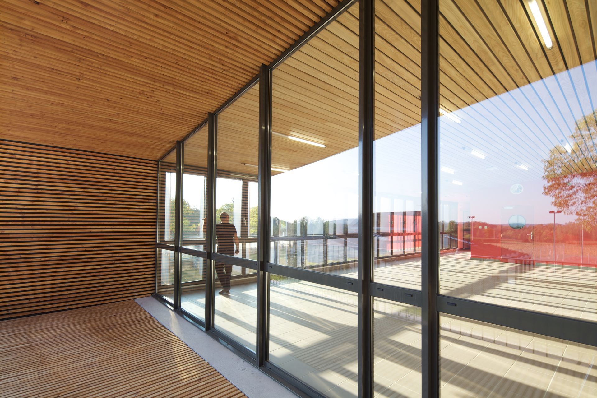 Vue intérieure : une personne se tient près de grandes baies vitrées donnant sur une clôture rouge et un espace extérieur. Éléments en bois.