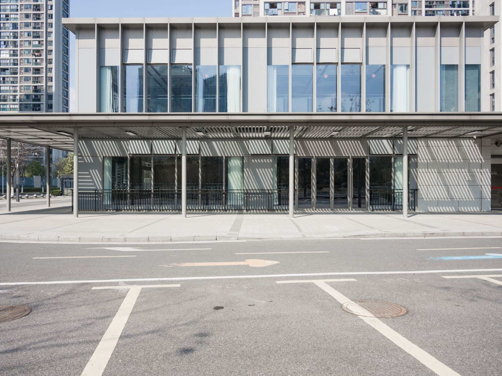 Bâtiment avec fenêtres en verre et entrée couverte, façade grise et blanche, parking au premier plan.