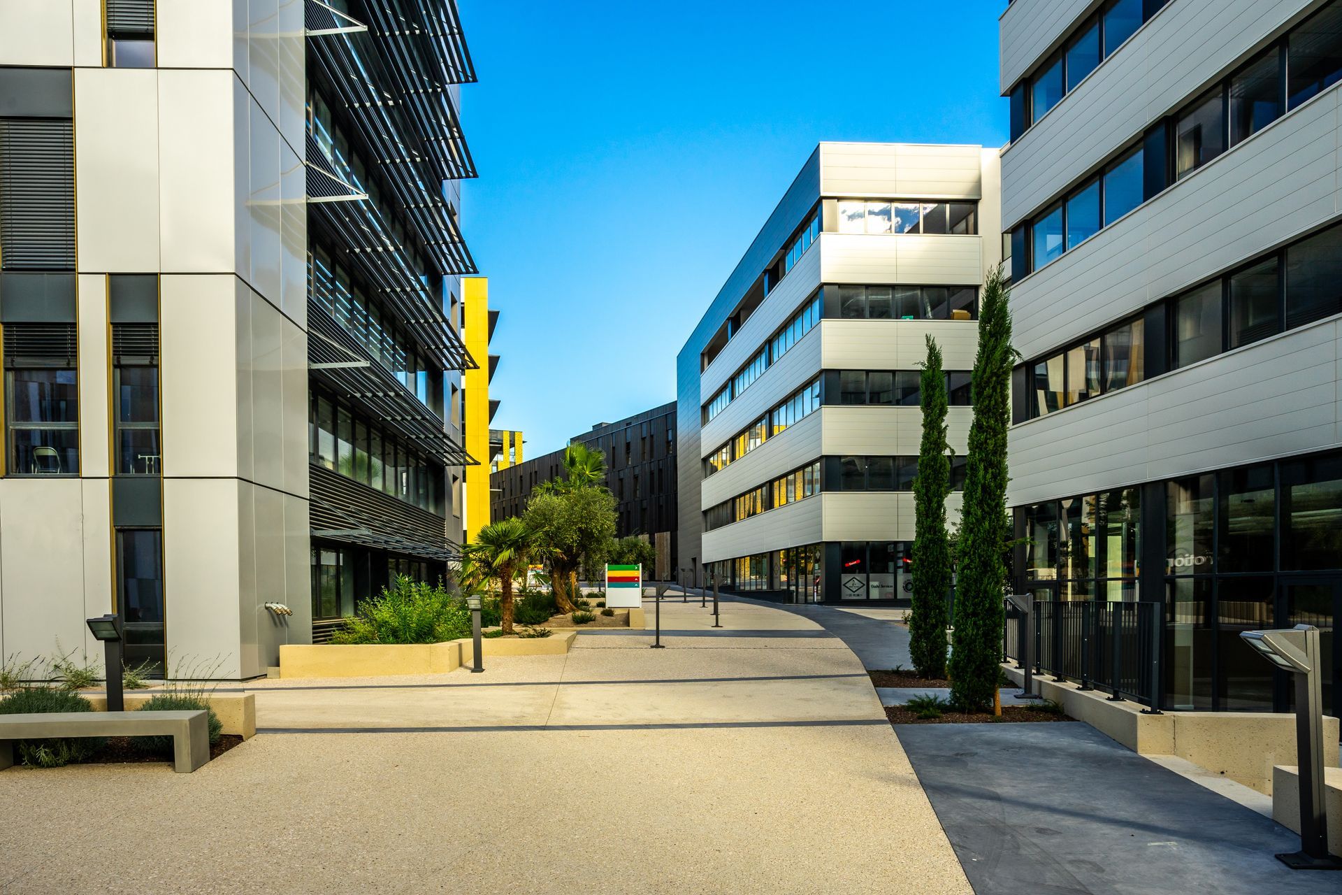 Des immeubles de bureaux modernes bordent une allée pavée sous un ciel bleu, agrémentée de quelques espaces verts.