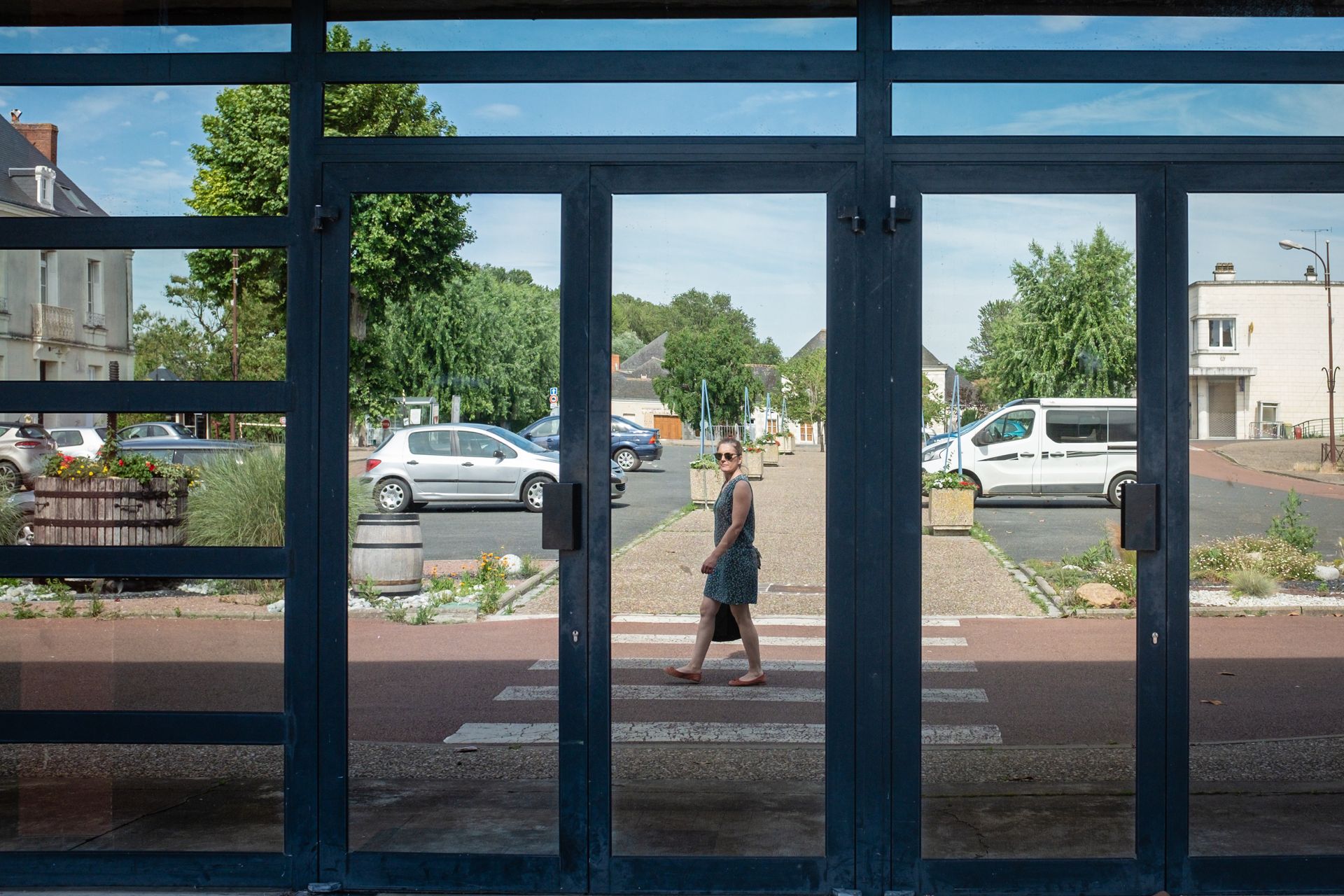Une femme traversant la rue se reflète dans des portes vitrées. On aperçoit des immeubles et des voitures.