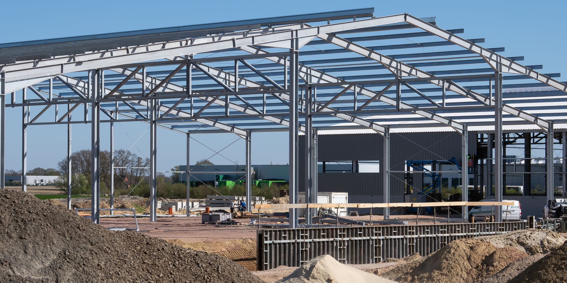 La structure métallique d'un bâtiment en construction, avec un tas de terre au premier plan et un ciel bleu au-dessus.