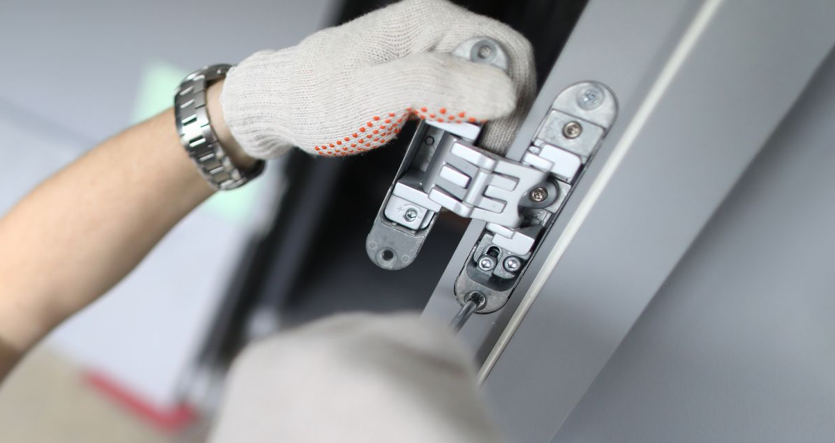 Main gantée ajustant une charnière métallique sur un cadre de porte gris.