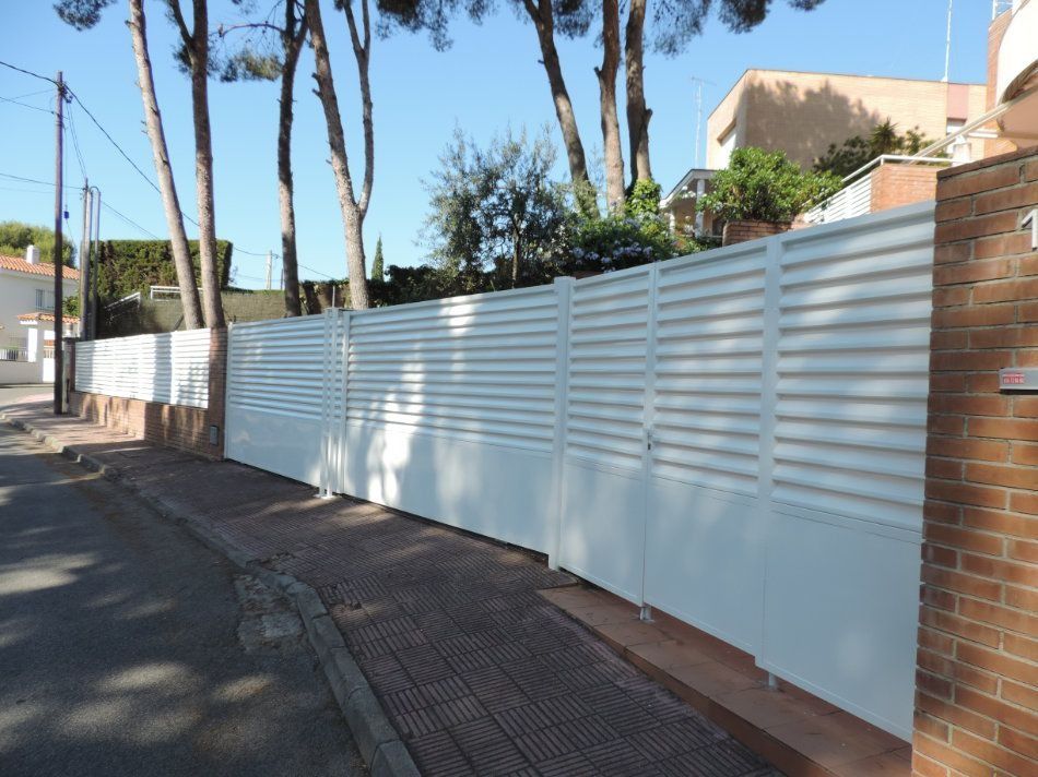 Una valla blanca con una pared de ladrillos y árboles al fondo.