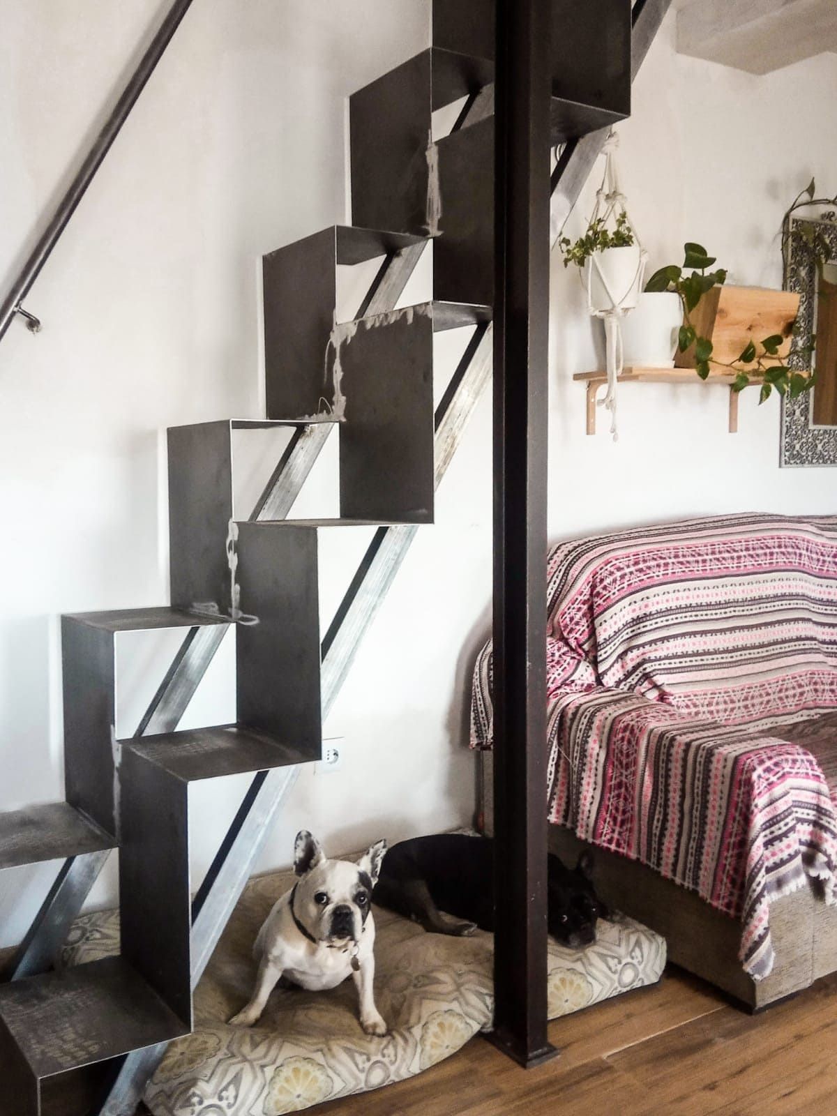 Un perro está sentado en una cama para perros debajo de una escalera en una sala de estar.