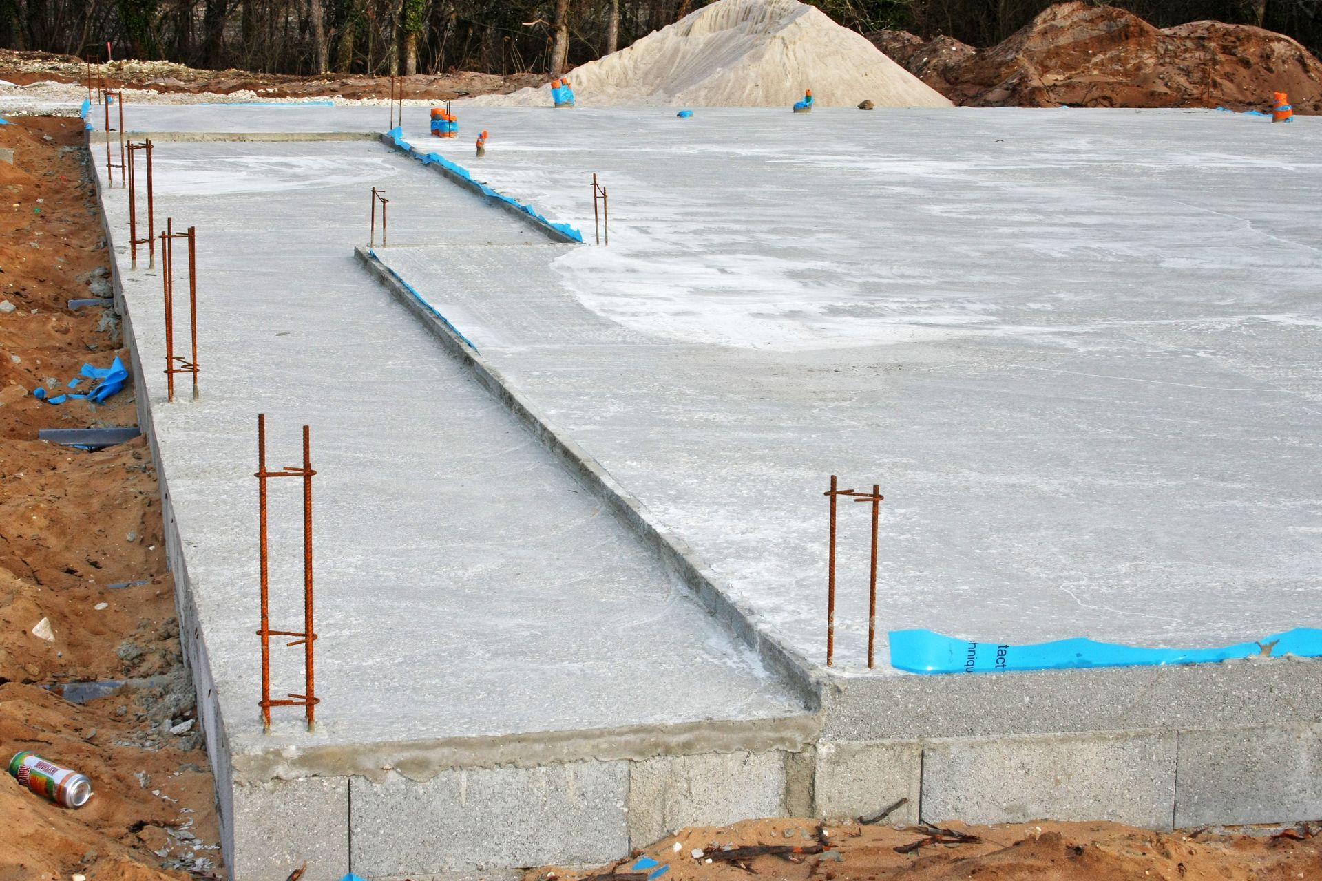 Fondations en béton d'un bâtiment en construction, avec des supports en barres d'armature et un tas de sable en arrière-plan.