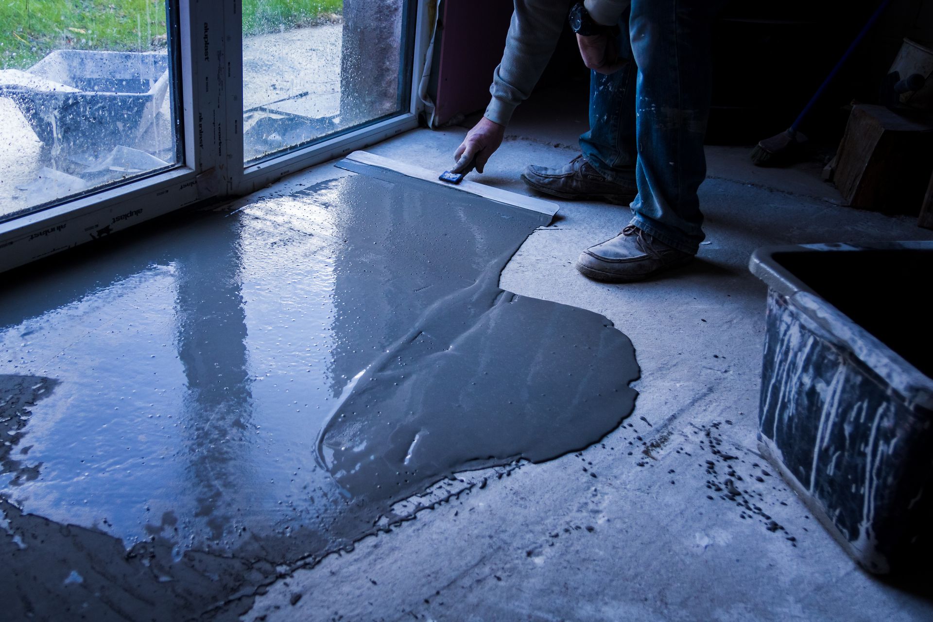 Personne lissant du béton sur un sol avec une truelle près d'une porte vitrée.