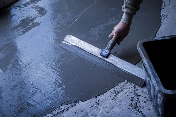 Une personne lisse du ciment humide sur un sol avec une truelle, à côté d'un seau foncé.
