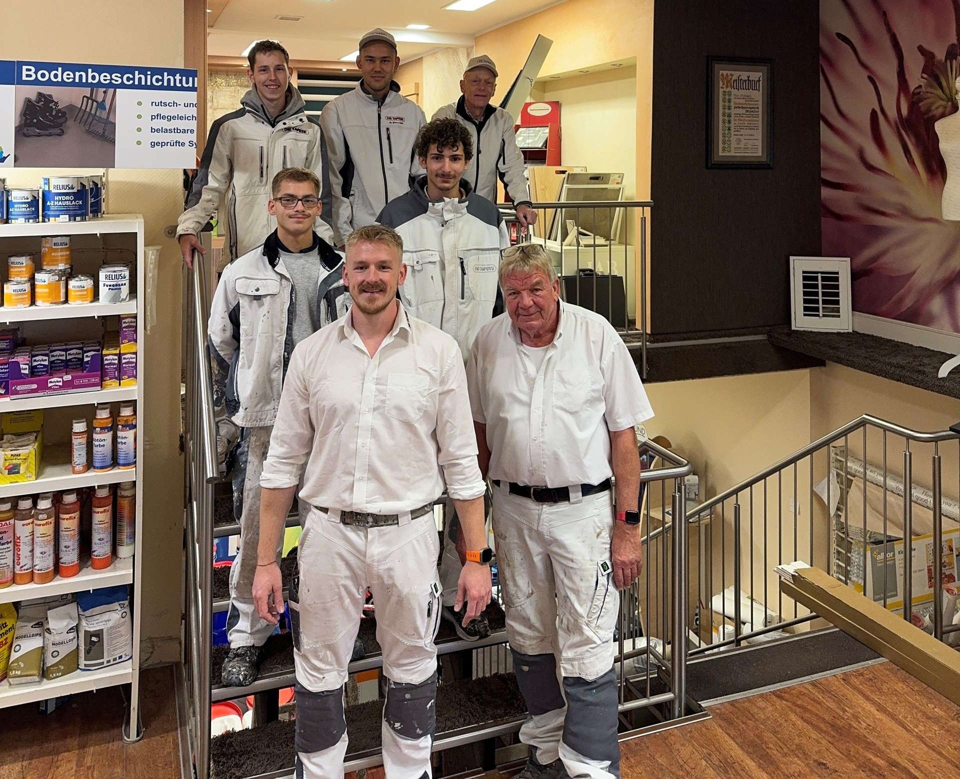Sieben Personen posieren in weißen Uniformen auf einer Treppe im Innenbereich, in einer Lobby oder einem Geschäft mit Regalen in der Nähe