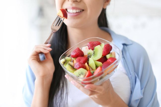 Mujer sonriendo, comiendo ensalada de frutas en un tazón de vidrio con un tenedor. Se ven fresas, kiwi y manzana.