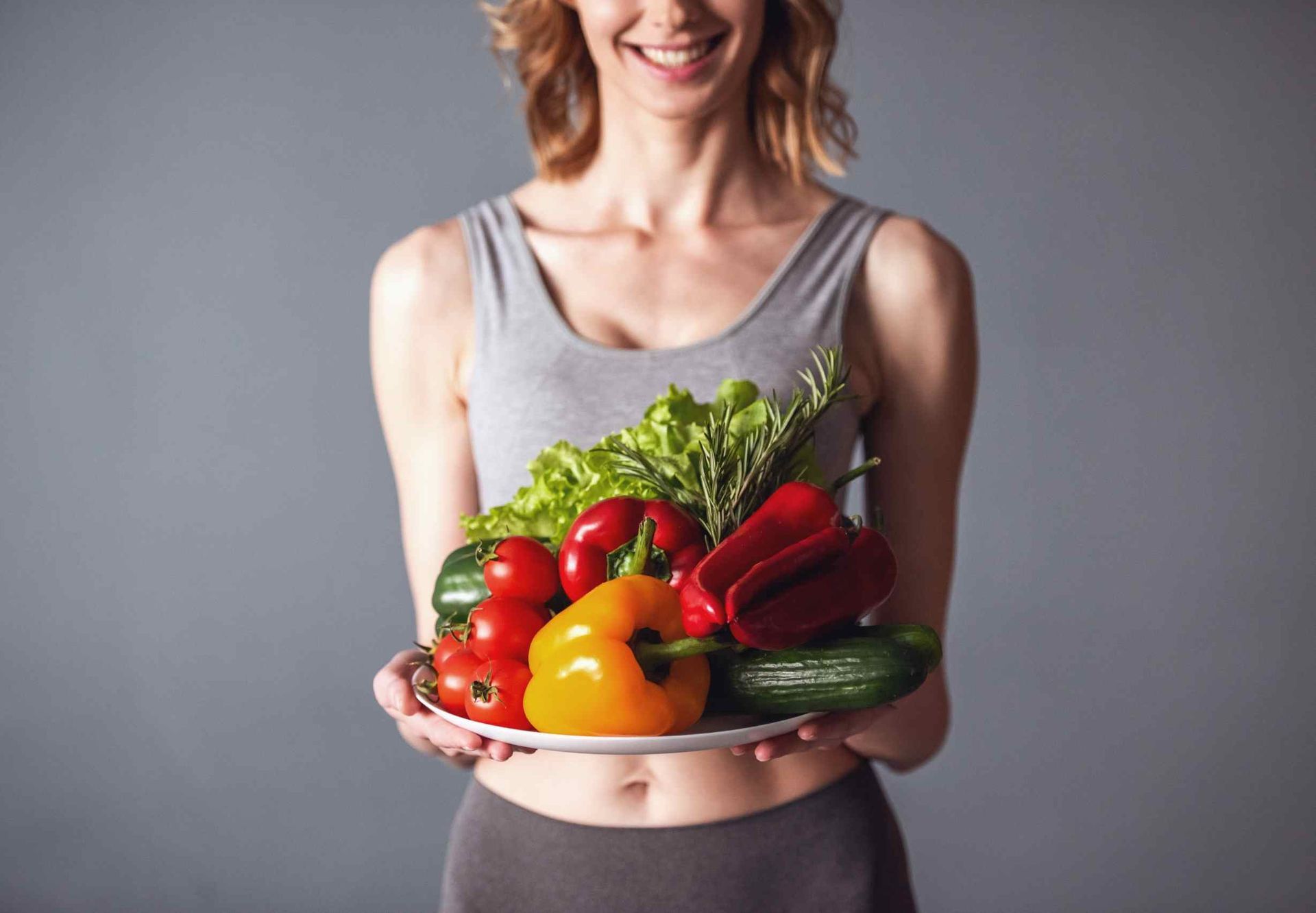 Mujer sonriendo, sosteniendo un plato de verduras frescas y coloridas sobre un fondo gris.