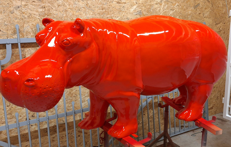 Sculpture d'hippopotame rouge vif derrière une clôture métallique adossée à un mur de pierre