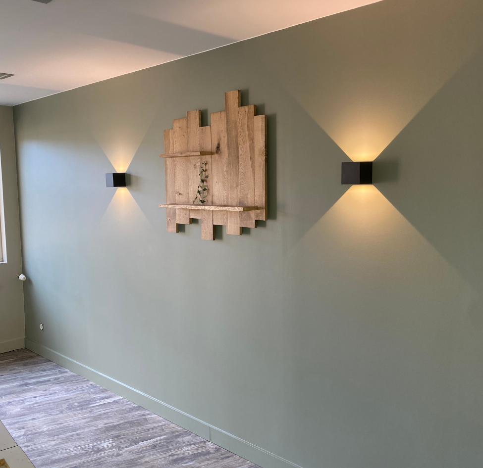 Mur peint en vert sauge orné d'une œuvre d'art géométrique en bois et de deux appliques noires diffusant une lumière douce.