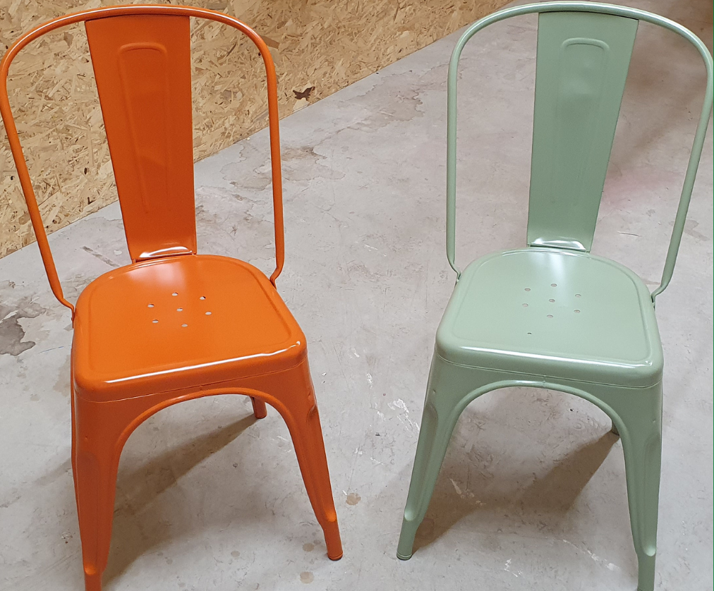 Deux chaises en métal, l'une orange et l'autre vert pâle, sur un sol en béton contre un mur beige.